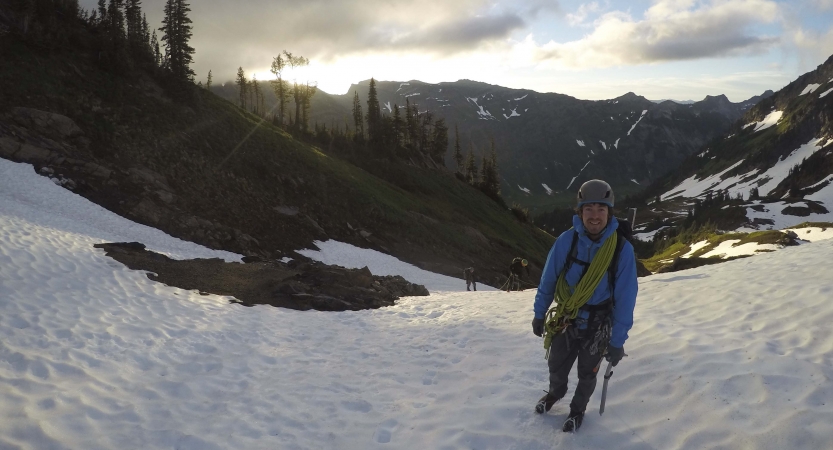 A person wearing safety gear stands on the snow in front of a vast mountainous landscape